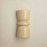 A minimalist, glossy ceramic wall sconce in a warm cream or pale yellow color, mounted on a plain beige wall.