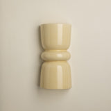 A minimalist, glossy ceramic wall sconce in a warm cream or pale yellow color, mounted on a plain beige wall.