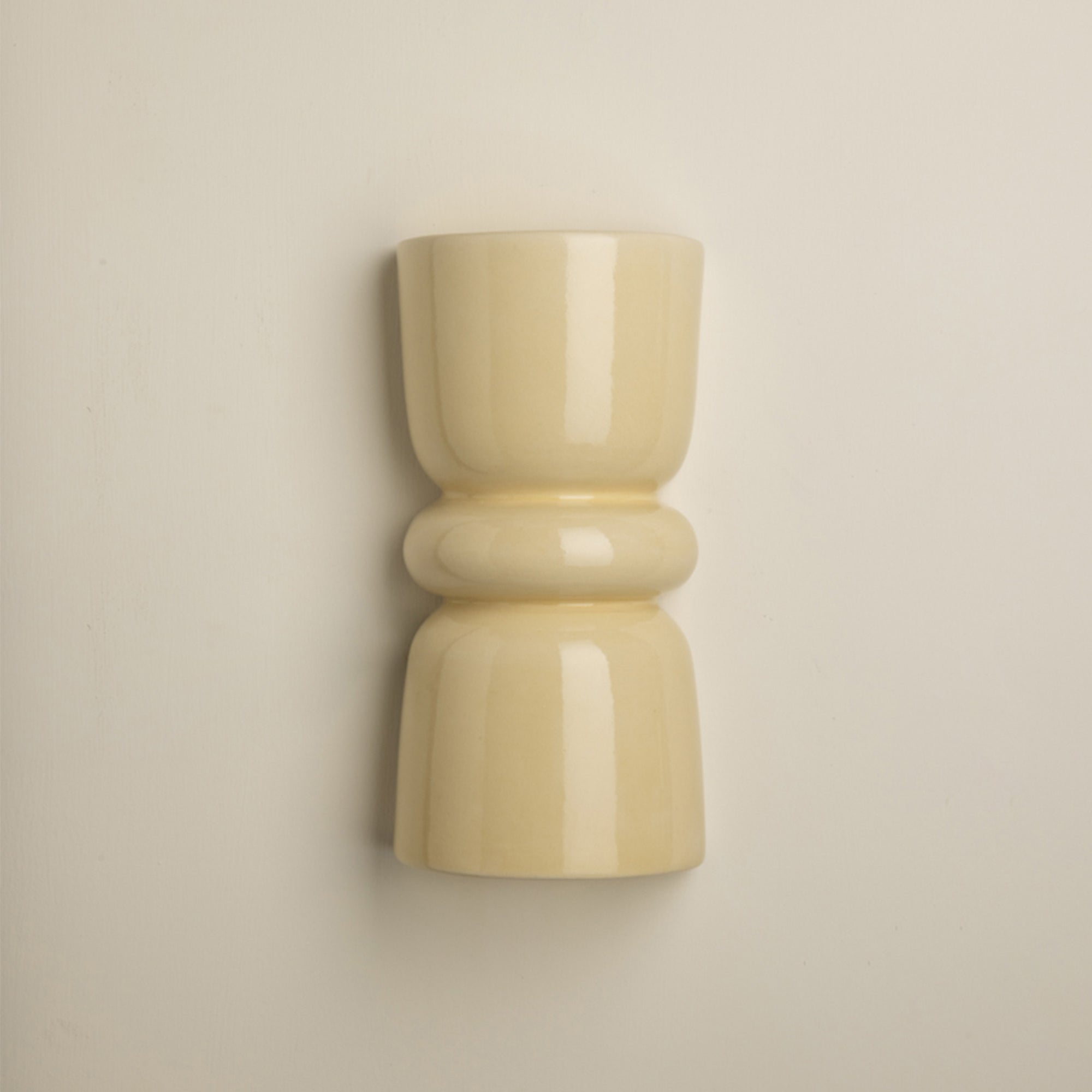 A minimalist, glossy ceramic wall sconce in a warm cream or pale yellow color, mounted on a plain beige wall.