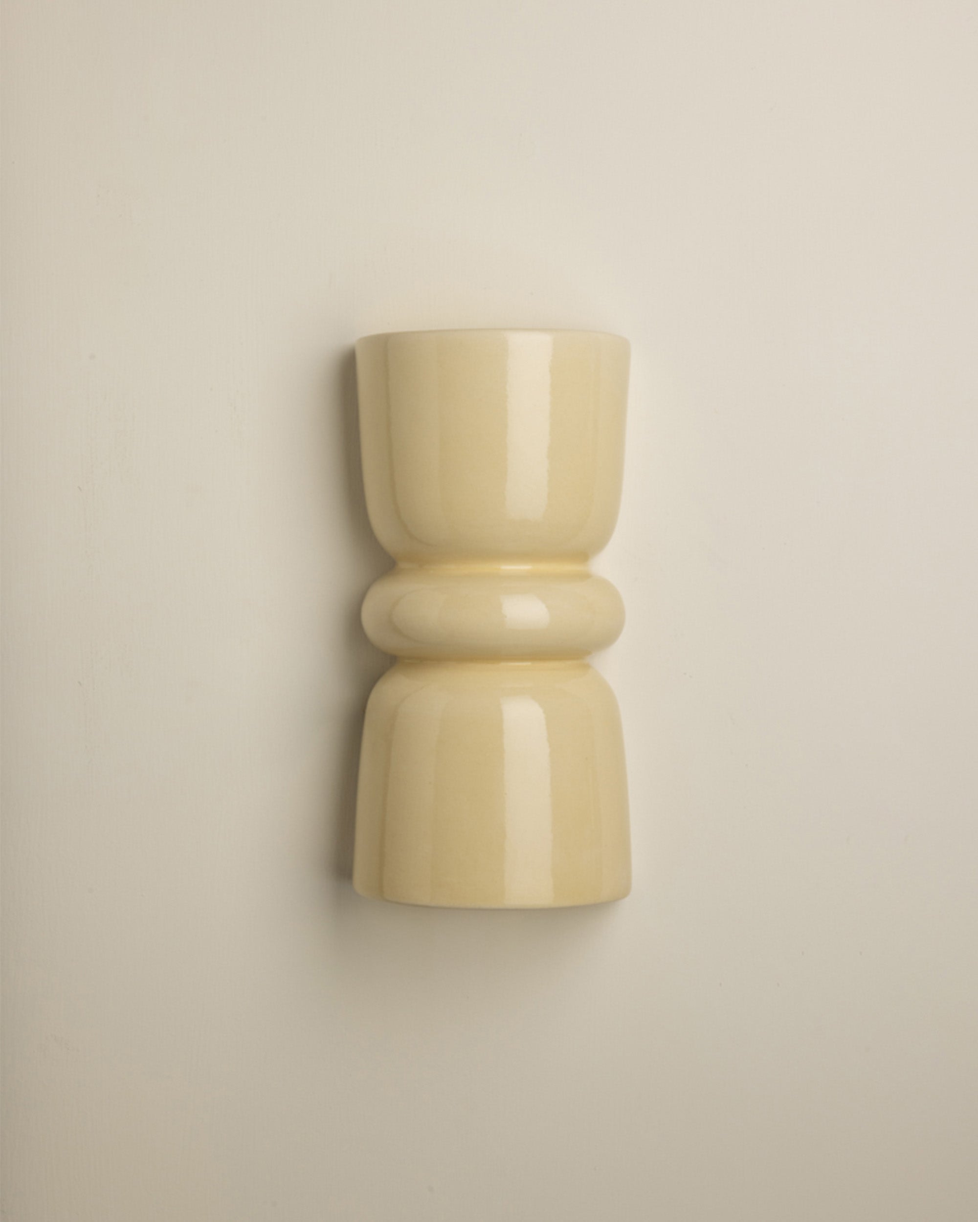 A minimalist, glossy ceramic wall sconce in a warm cream or pale yellow color, mounted on a plain beige wall.