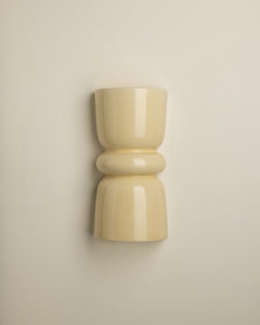 A minimalist, glossy ceramic wall sconce in a warm cream or pale yellow color, mounted on a plain beige wall.