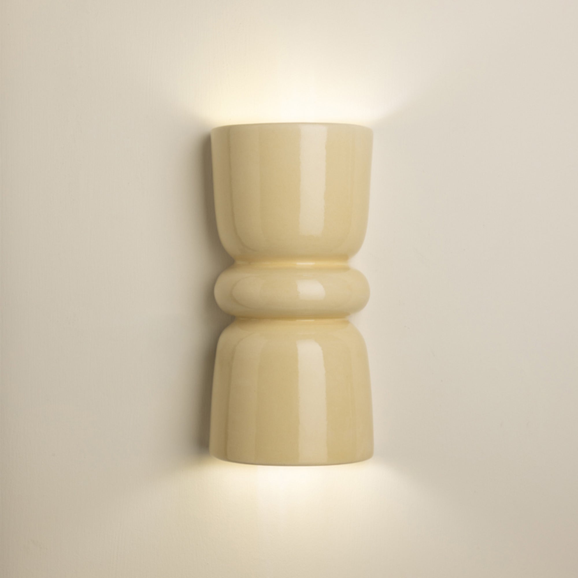 A modern wall-mounted sconce with a glossy, rounded cylindrical design in a warm cream or beige finish, emitting a soft warm light against a neutral beige wall.