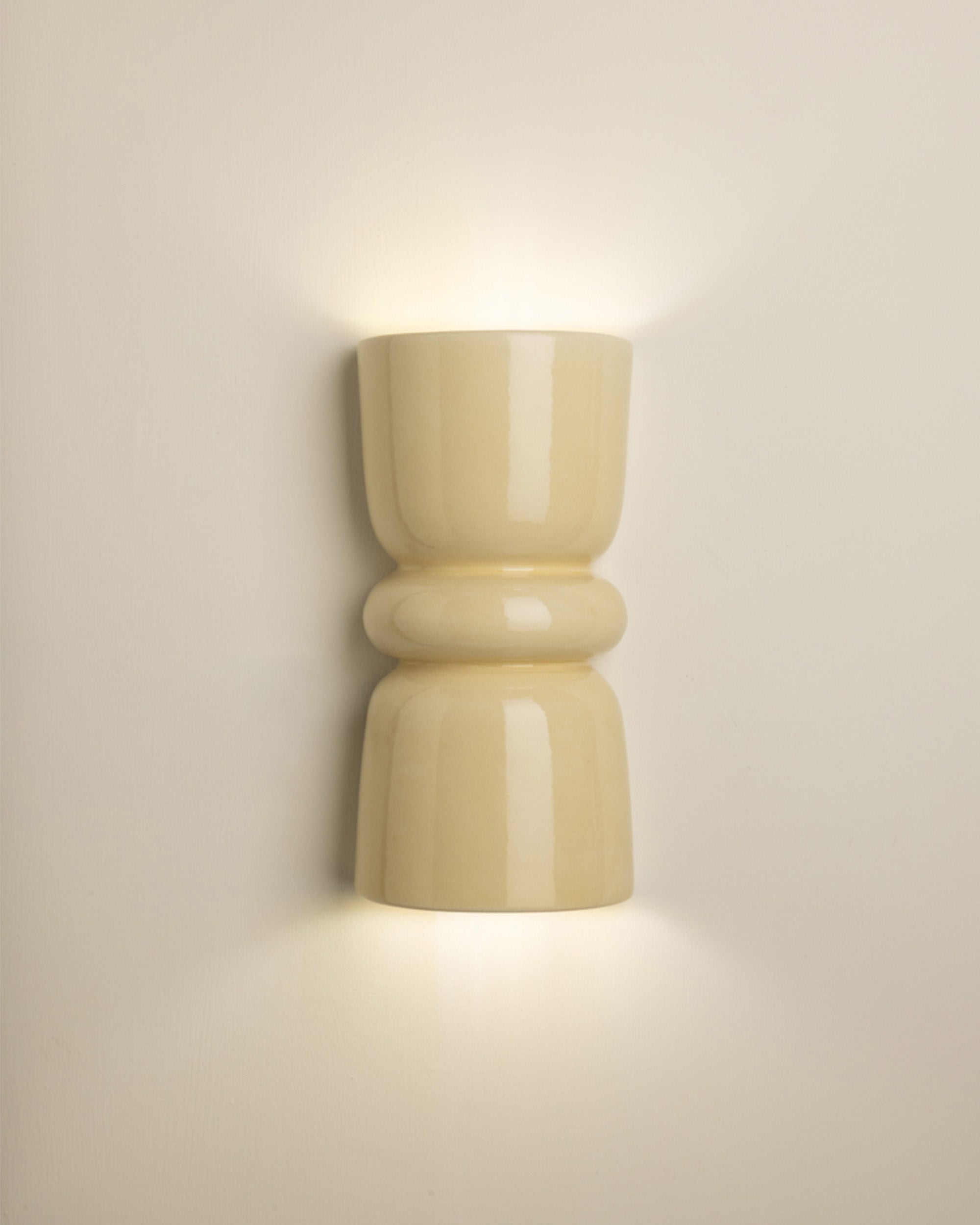 A modern wall-mounted sconce with a glossy, rounded cylindrical design in a warm cream or beige finish, emitting a soft warm light against a neutral beige wall.