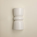 A minimalist white ceramic wall sconce with a sculptural, hourglass-like shape and rounded bands, mounted on a warm beige wall.