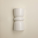 A minimalist white ceramic wall sconce with a sculptural, hourglass-like shape and rounded bands, mounted on a warm beige wall.