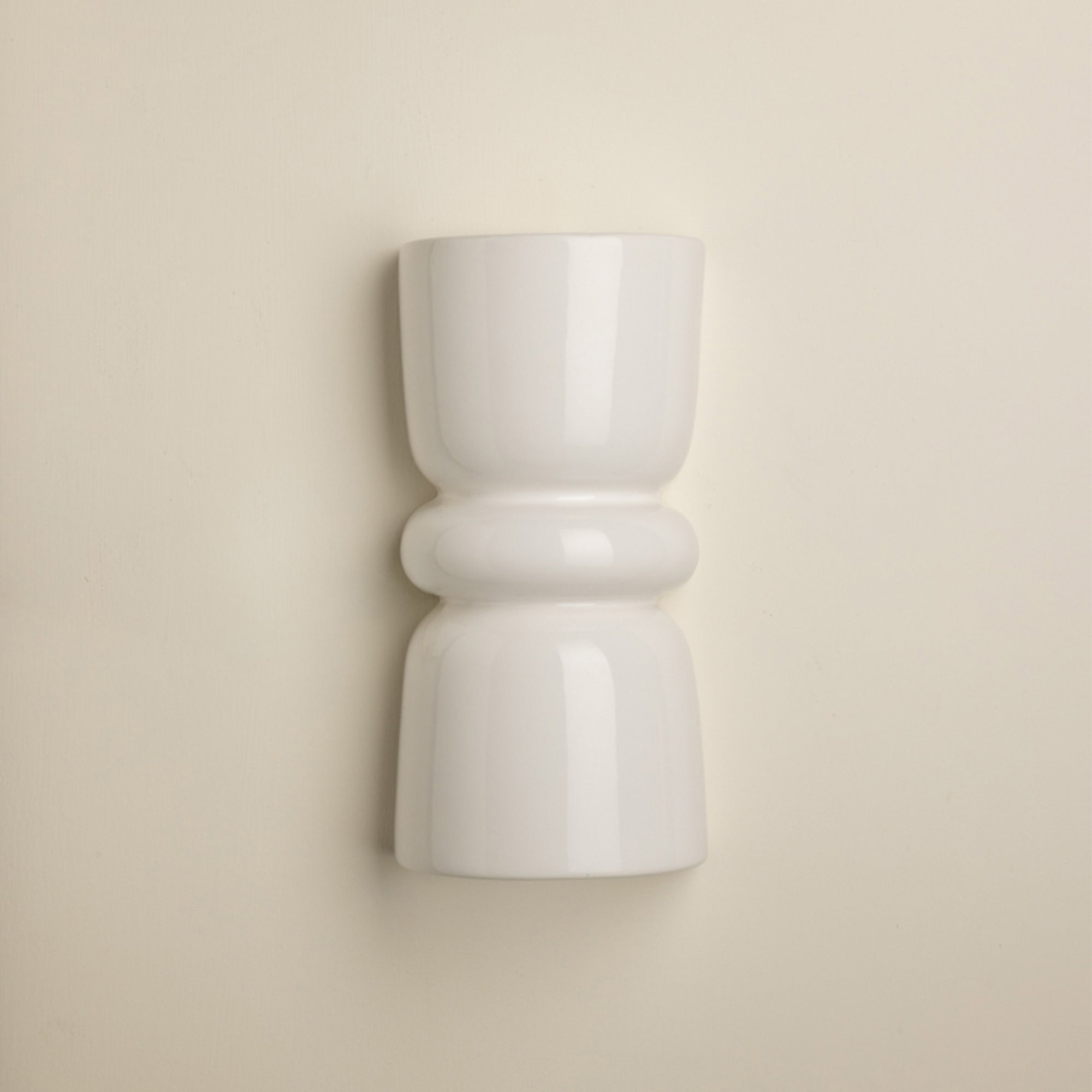 A minimalist white ceramic wall sconce with a sculptural, hourglass-like shape and rounded bands, mounted on a warm beige wall.