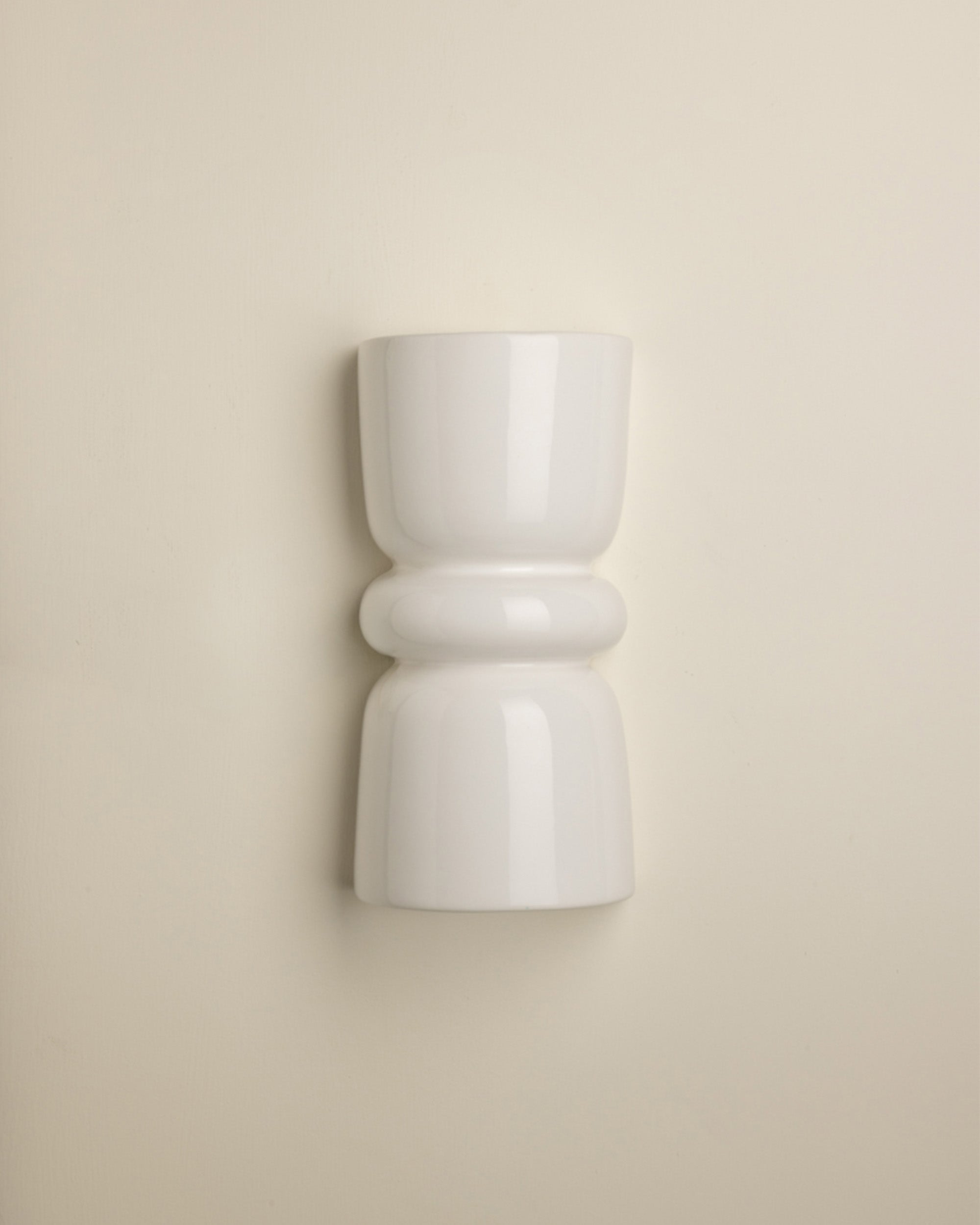 A minimalist white ceramic wall sconce with a sculptural, hourglass-like shape and rounded bands, mounted on a warm beige wall.