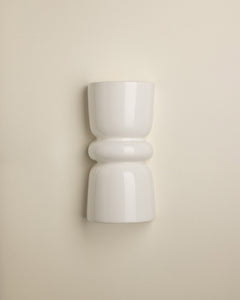 A minimalist white ceramic wall sconce with a sculptural, hourglass-like shape and rounded bands, mounted on a warm beige wall.