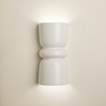 A modern white ceramic wall sconce with a sculptural hourglass shape, shown illuminated against a warm beige wall.