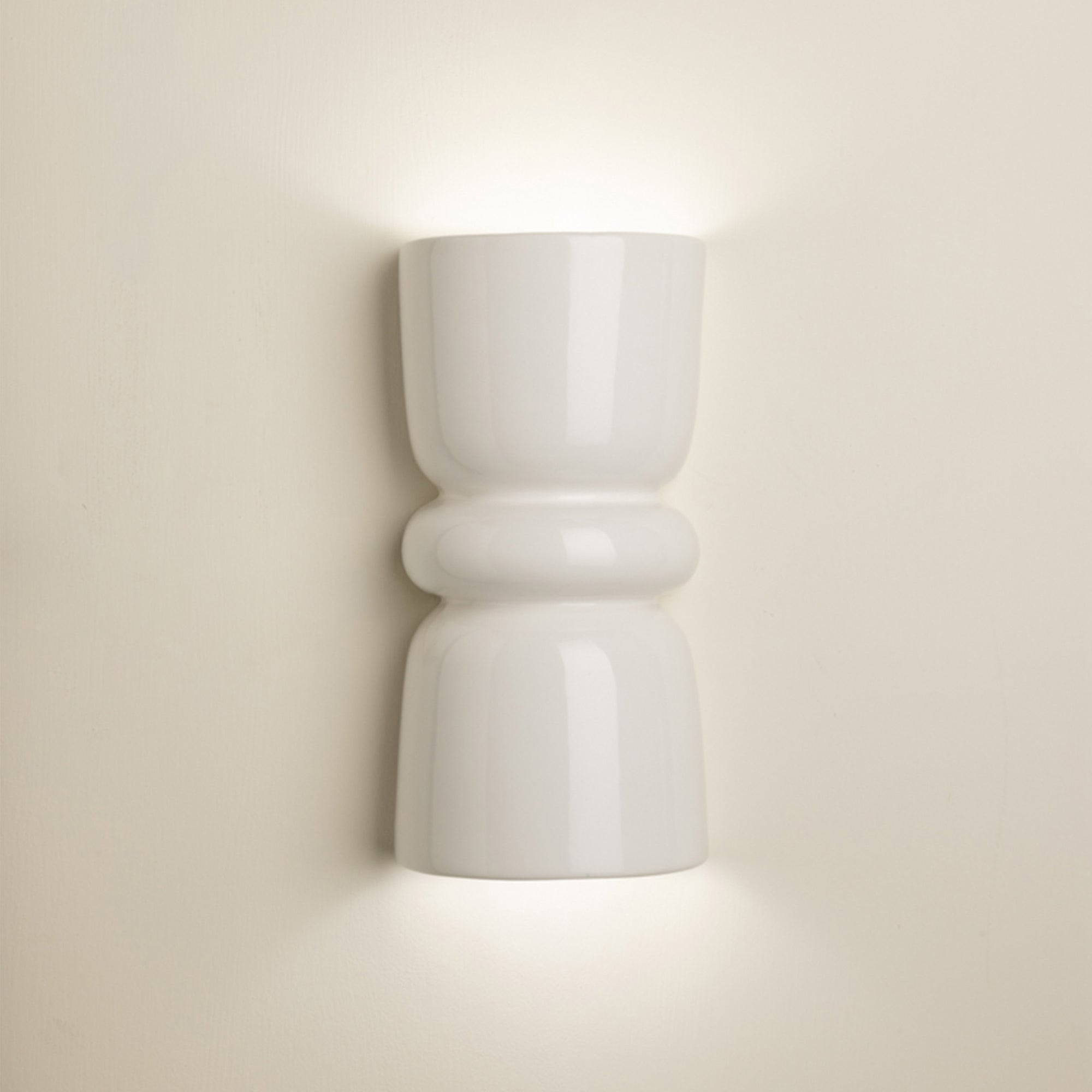 A modern white ceramic wall sconce with a sculptural hourglass shape, shown illuminated against a warm beige wall.