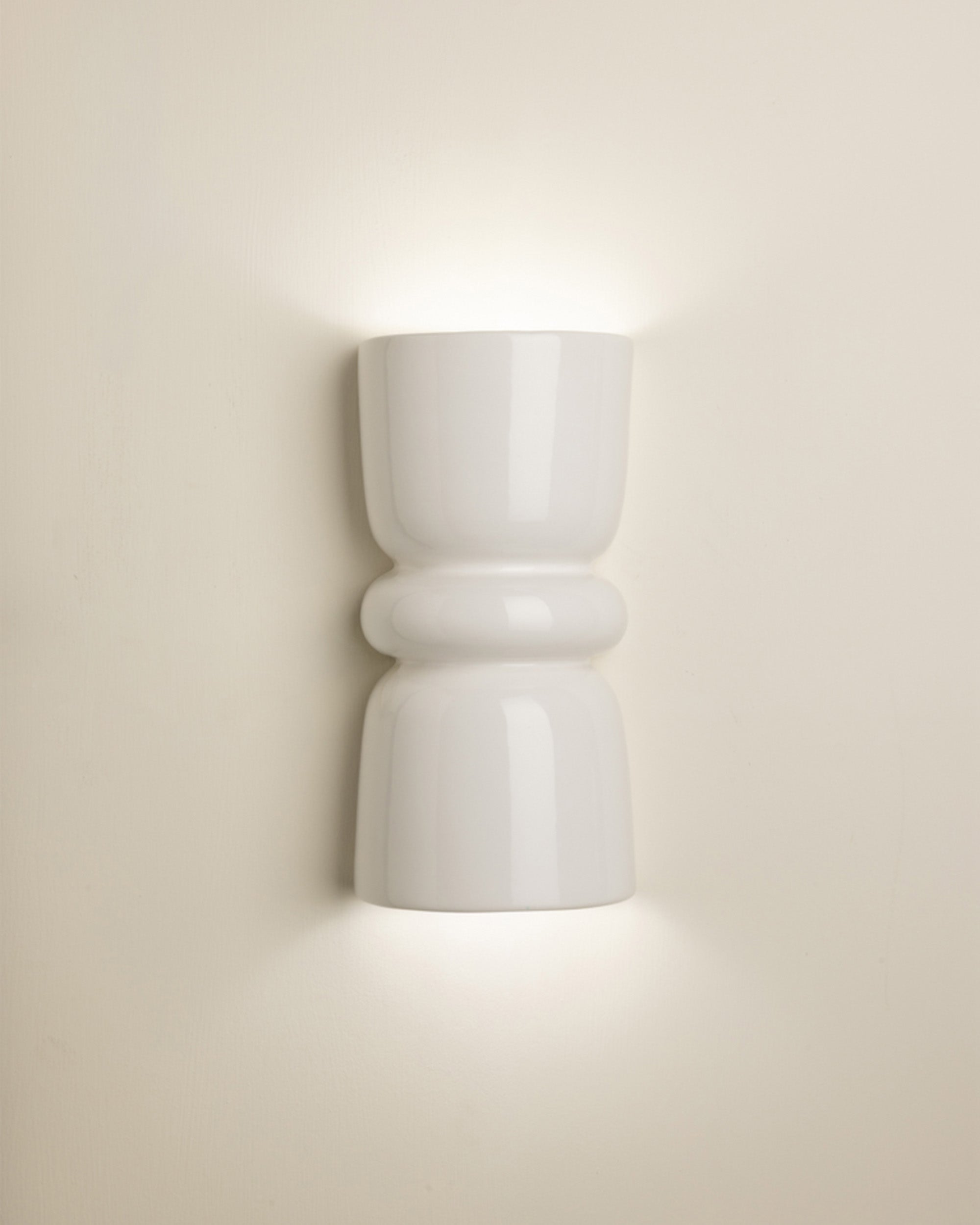 A modern white ceramic wall sconce with a sculptural hourglass shape, shown illuminated against a warm beige wall.