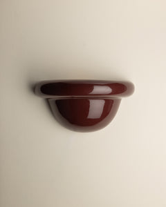 A glossy, dark reddish-brown wall-mounted shelf or hook with a rounded, semi-circular profile, shown against a plain off-white background.