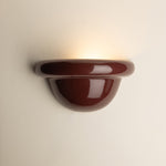 A glossy burgundy ceramic wall sconce light fixture mounted on a plain off-white wall, casting a warm yellow light upward.