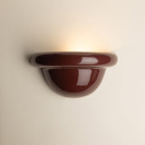 A glossy burgundy ceramic wall sconce light fixture mounted on a plain off-white wall, casting a warm yellow light upward.