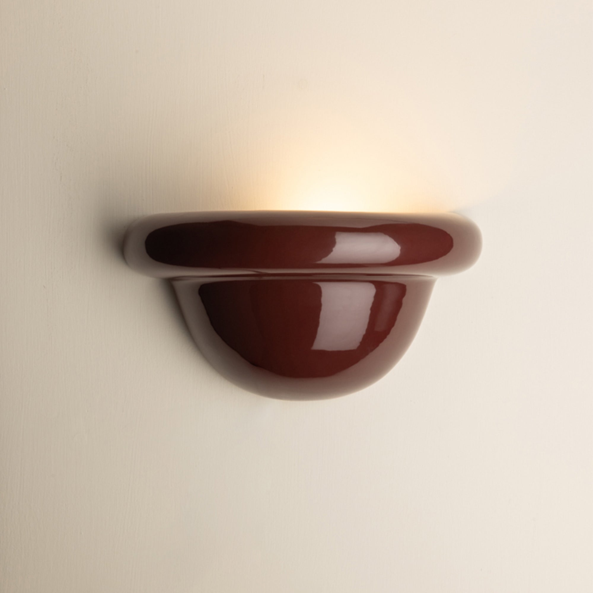 A glossy burgundy ceramic wall sconce light fixture mounted on a plain off-white wall, casting a warm yellow light upward.
