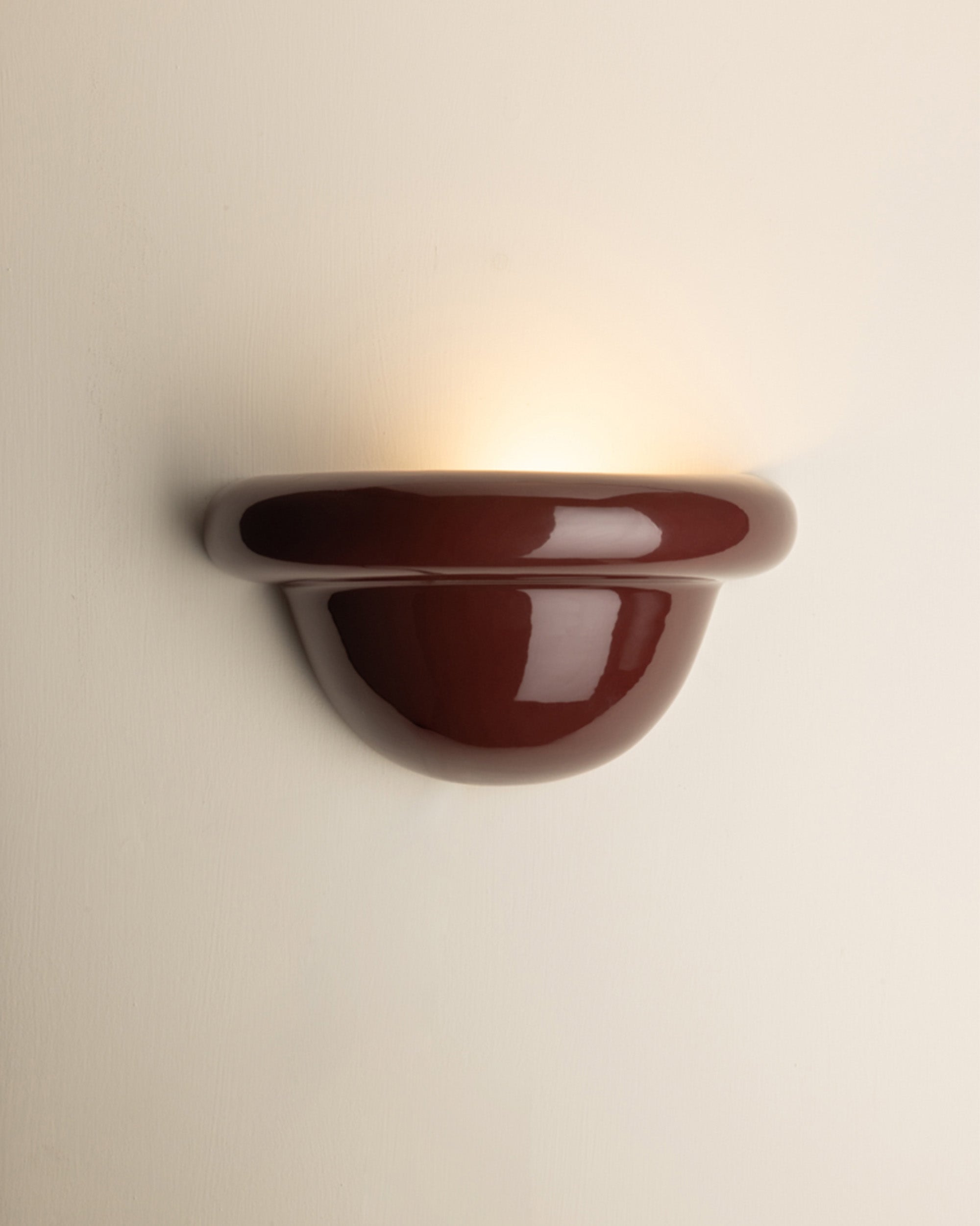 A glossy burgundy ceramic wall sconce light fixture mounted on a plain off-white wall, casting a warm yellow light upward.