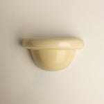 A glossy, dome-shaped wall-mounted sconce in a pale cream or butter yellow color, shown against a plain, light beige wall.