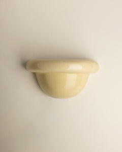 A glossy, dome-shaped wall-mounted sconce in a pale cream or butter yellow color, shown against a plain, light beige wall.