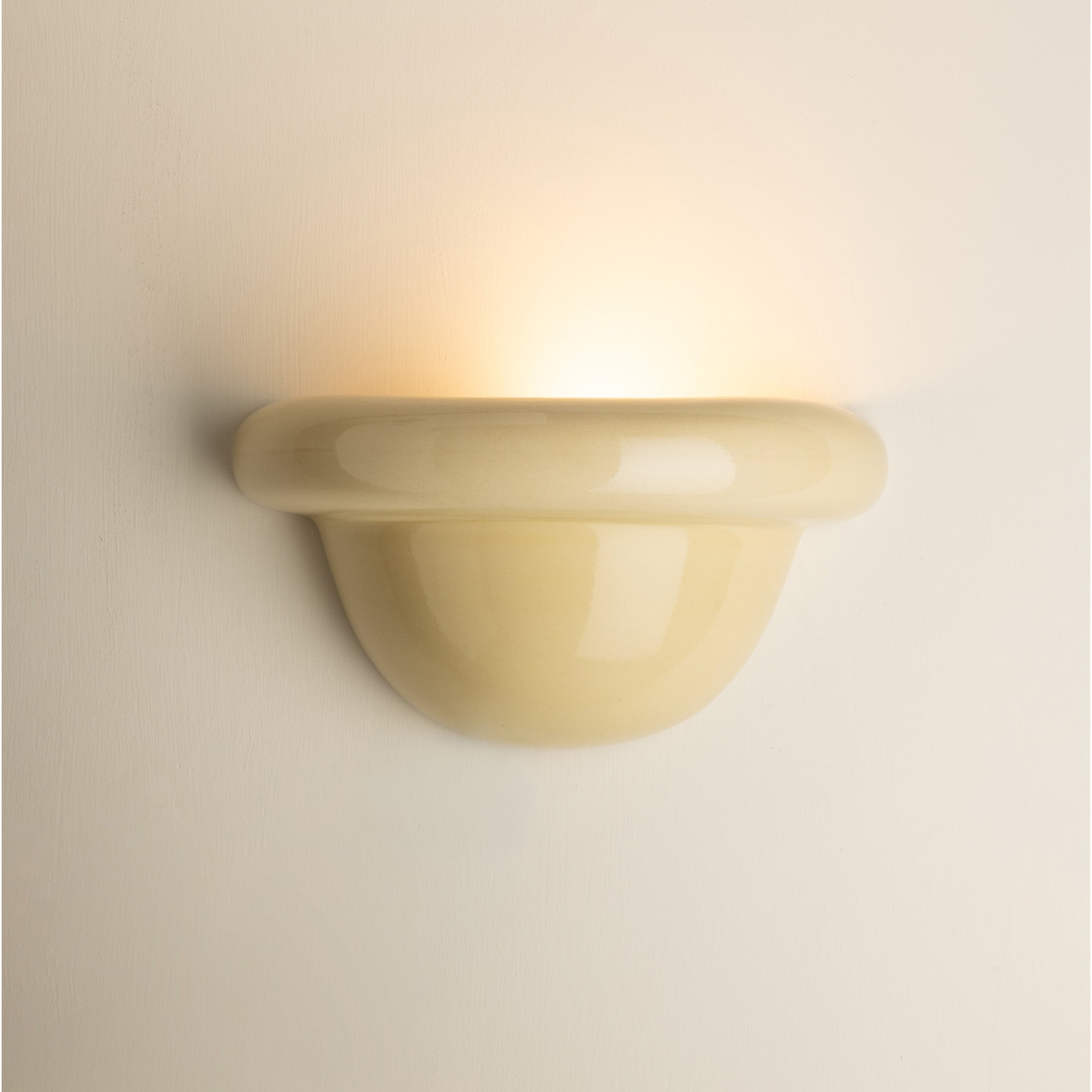 A modern mushroom-shaped wall sconce in a glossy cream finish, emitting a warm yellow light against a matching cream-colored wall.