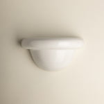 A minimalist white wall-mounted light fixture with a smooth, curved dome and a thick horizontal band, shown against a solid, warm off-white background.