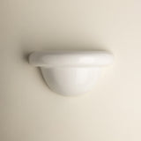 A minimalist white wall-mounted light fixture with a smooth, curved dome and a thick horizontal band, shown against a solid, warm off-white background.