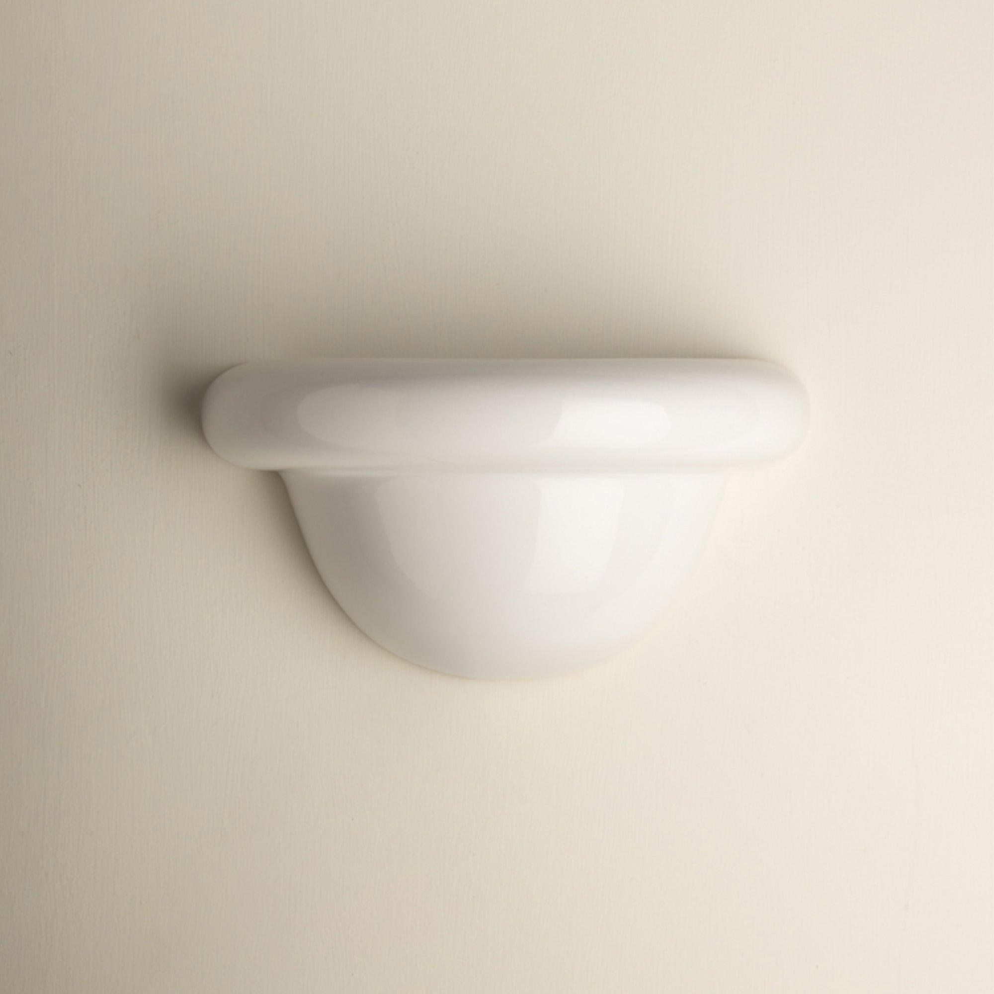 A minimalist white wall-mounted light fixture with a smooth, curved dome and a thick horizontal band, shown against a solid, warm off-white background.