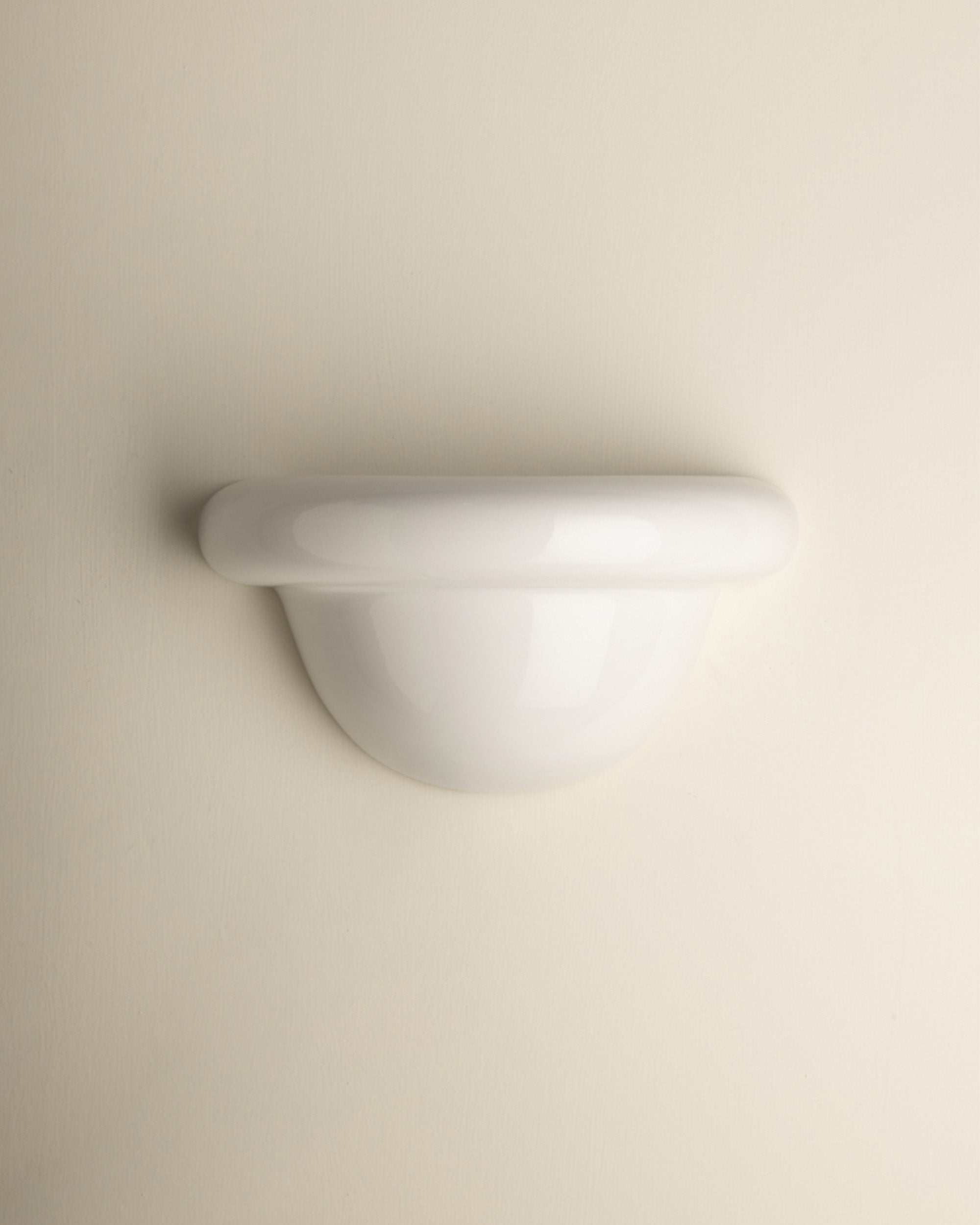 A minimalist white wall-mounted light fixture with a smooth, curved dome and a thick horizontal band, shown against a solid, warm off-white background.