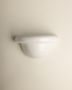 A minimalist white wall-mounted light fixture with a smooth, curved dome and a thick horizontal band, shown against a solid, warm off-white background.