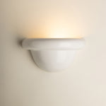 A modern white ceramic wall sconce light fixture mounted on a warm beige wall, casting a soft warm yellow glow upwards.