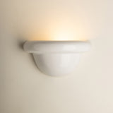 A modern white ceramic wall sconce light fixture mounted on a warm beige wall, casting a soft warm yellow glow upwards.