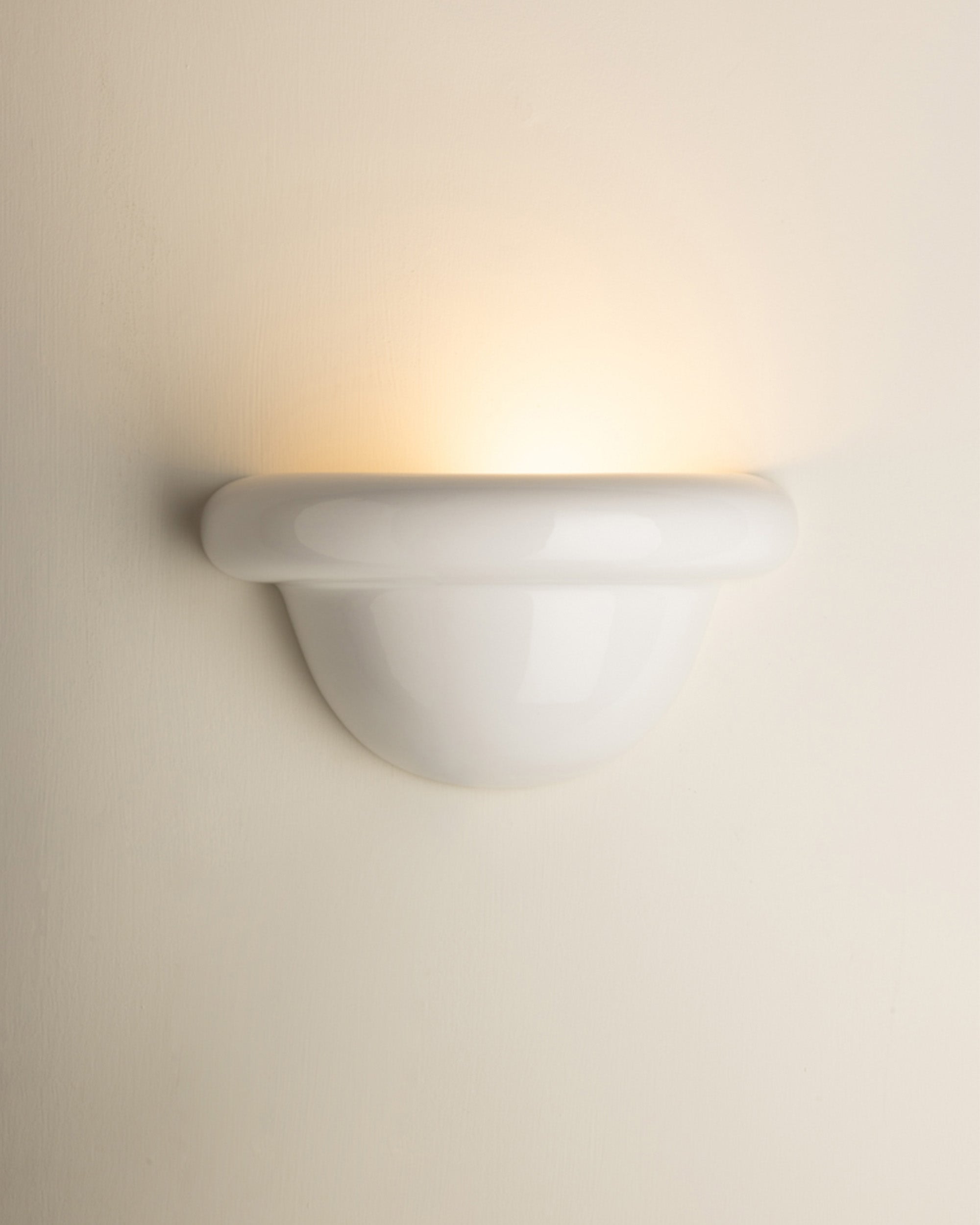 A modern white ceramic wall sconce light fixture mounted on a warm beige wall, casting a soft warm yellow glow upwards.