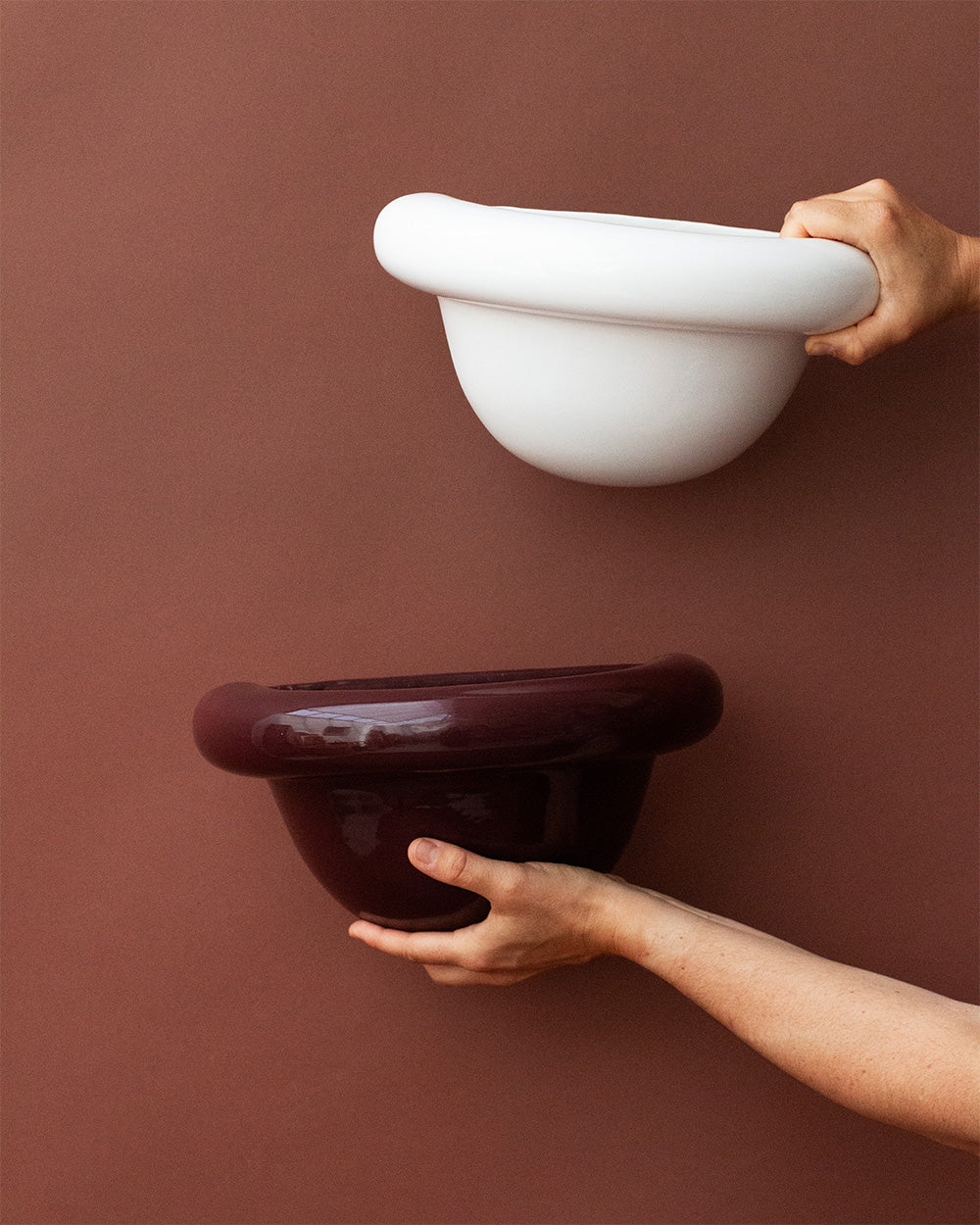Two hands holding a white and a dark brown ceramic wall sconce against a brown background