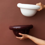 Two hands holding a white and a dark brown ceramic wall sconce against a brown background