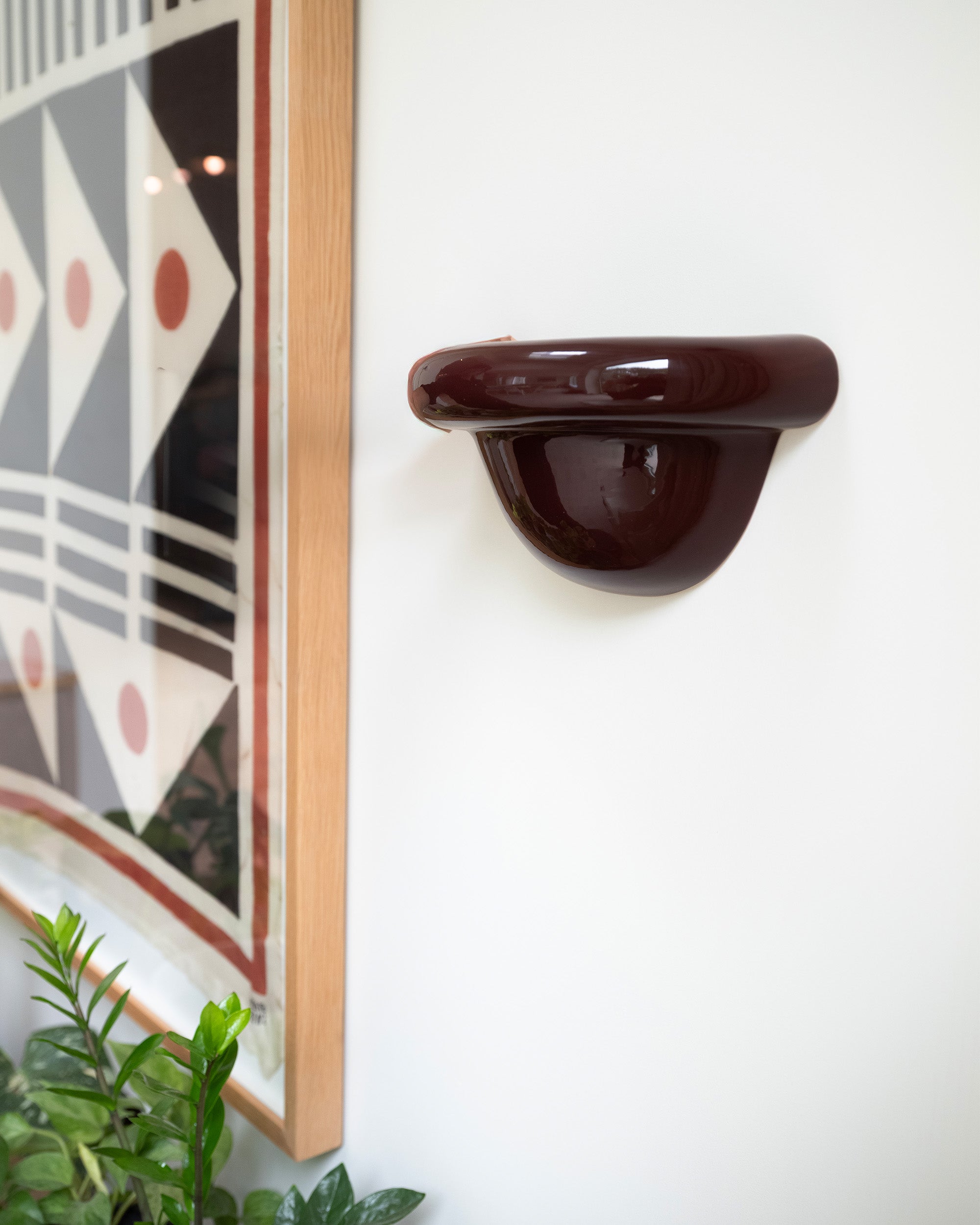 Late Afternoon Lighting Burgundy Uplighter Ceramic Sconce Wall Light hung on a white wall next to a framed print
