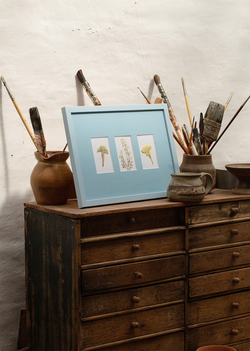 A light blue framed art print featuring three small botanical illustrations, styled on top of a rustic wooden chest of drawers with paintbrushes and pottery against a textured white wall.