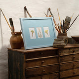 A light blue framed art print featuring three small botanical illustrations, styled on top of a rustic wooden chest of drawers with paintbrushes and pottery against a textured white wall.