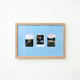 A light wood framed wall art piece with a Sky blue mount and three small vertical landscape photographs arranged side by side, mounted on a plain white wall.
