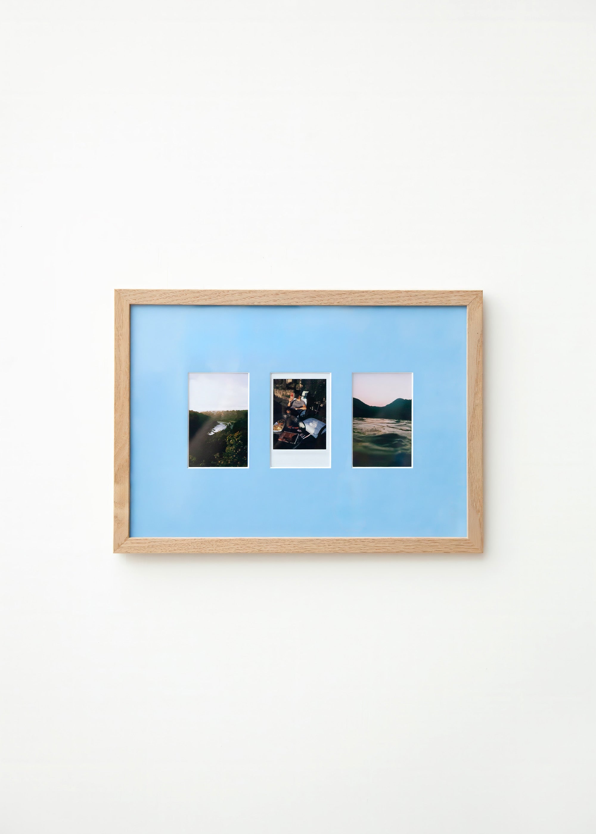 A light wood framed wall art piece with a Sky blue mount and three small vertical landscape photographs arranged side by side, mounted on a plain white wall.