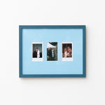 A blue picture frame with a light blue mat, holding three vertical instant-style photographs, shown hanging on a plain white wall.