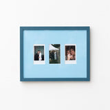 A blue picture frame with a light blue mat, holding three vertical instant-style photographs, shown hanging on a plain white wall.