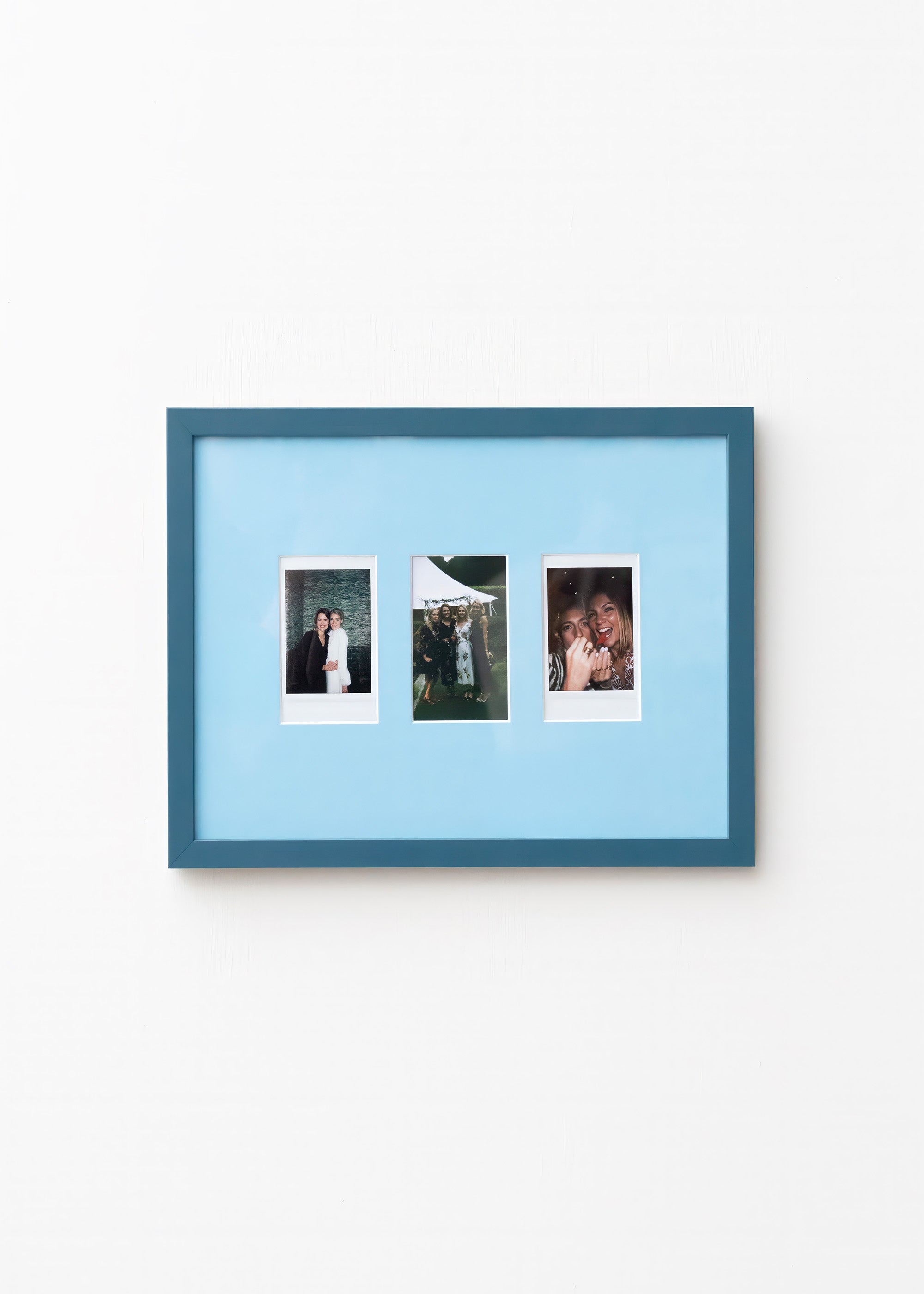 A blue picture frame with a light blue mat, holding three vertical instant-style photographs, shown hanging on a plain white wall.