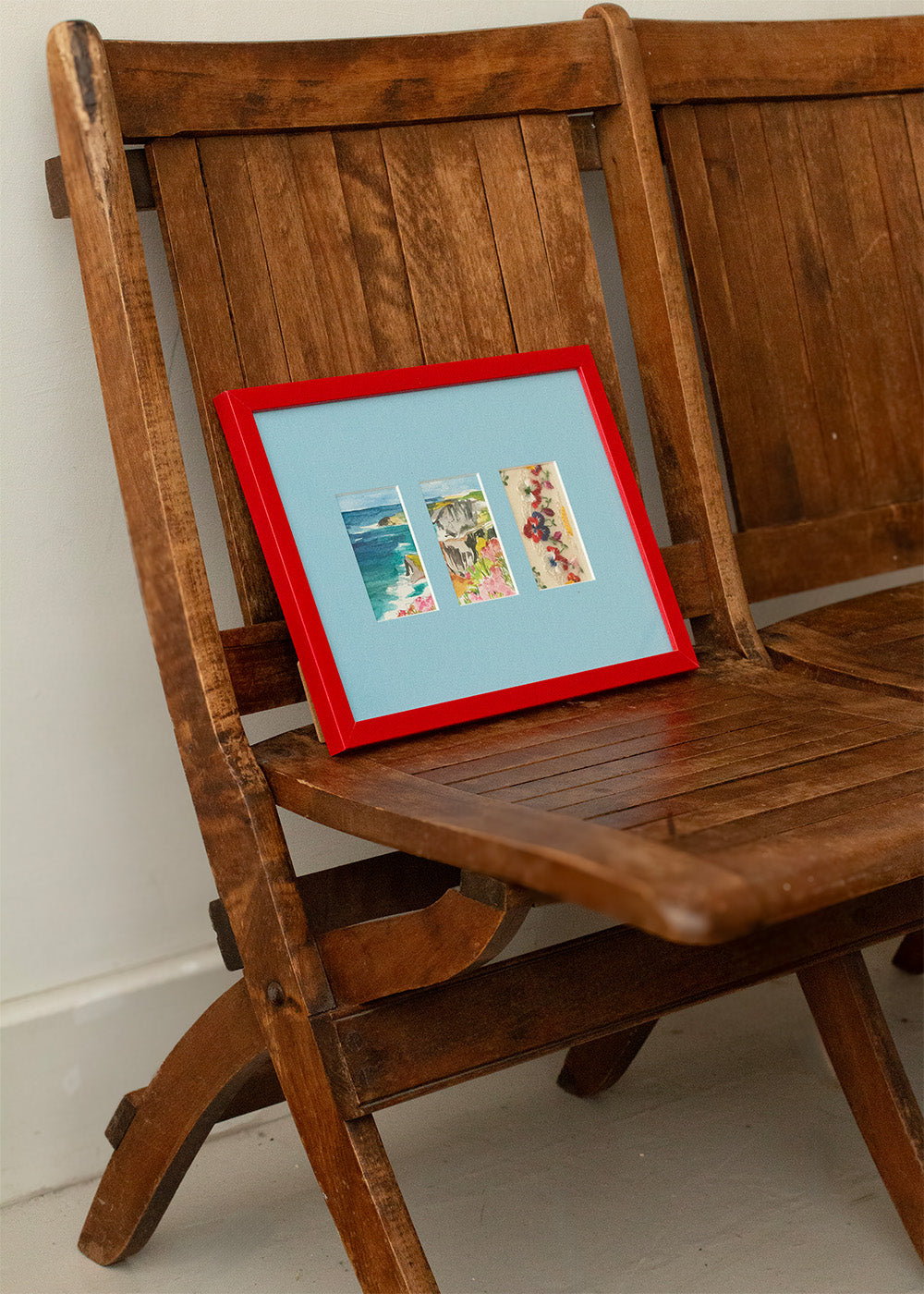 A red-framed picture frame with three colourful beach-themed postcards on a light blue mount, leaning on a rustic wooden chair.