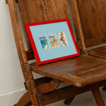 A red-framed picture frame with three colourful beach-themed postcards on a light blue mount, leaning on a rustic wooden chair.