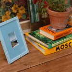 A light blue wooden picture frame with a small inset photograph, styled on a rustic wooden table next to a stack of books and a potted plant.