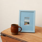 A light blue picture frame with a small landscape photograph inside, styled on a wooden table next to a brown ceramic mug against a plain off-white wall.