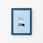A dark blue picture frame with a light blue mat, displaying a small landscape photograph of mountains and yellow skis, set against a plain white wall.