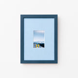 A dark blue picture frame with a light blue mat, displaying a small landscape photograph of mountains and yellow skis, set against a plain white wall.