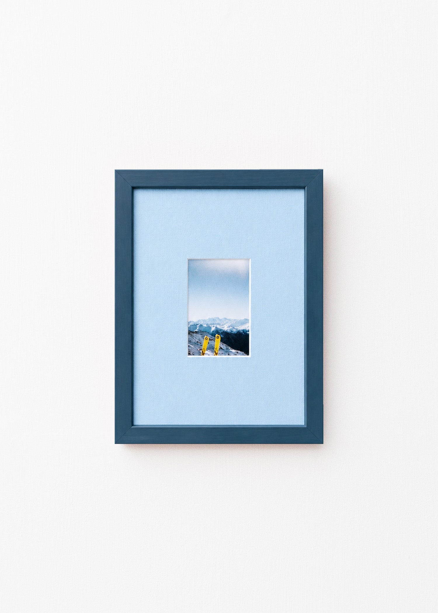 A dark blue picture frame with a light blue mat, displaying a small landscape photograph of mountains and yellow skis, set against a plain white wall.