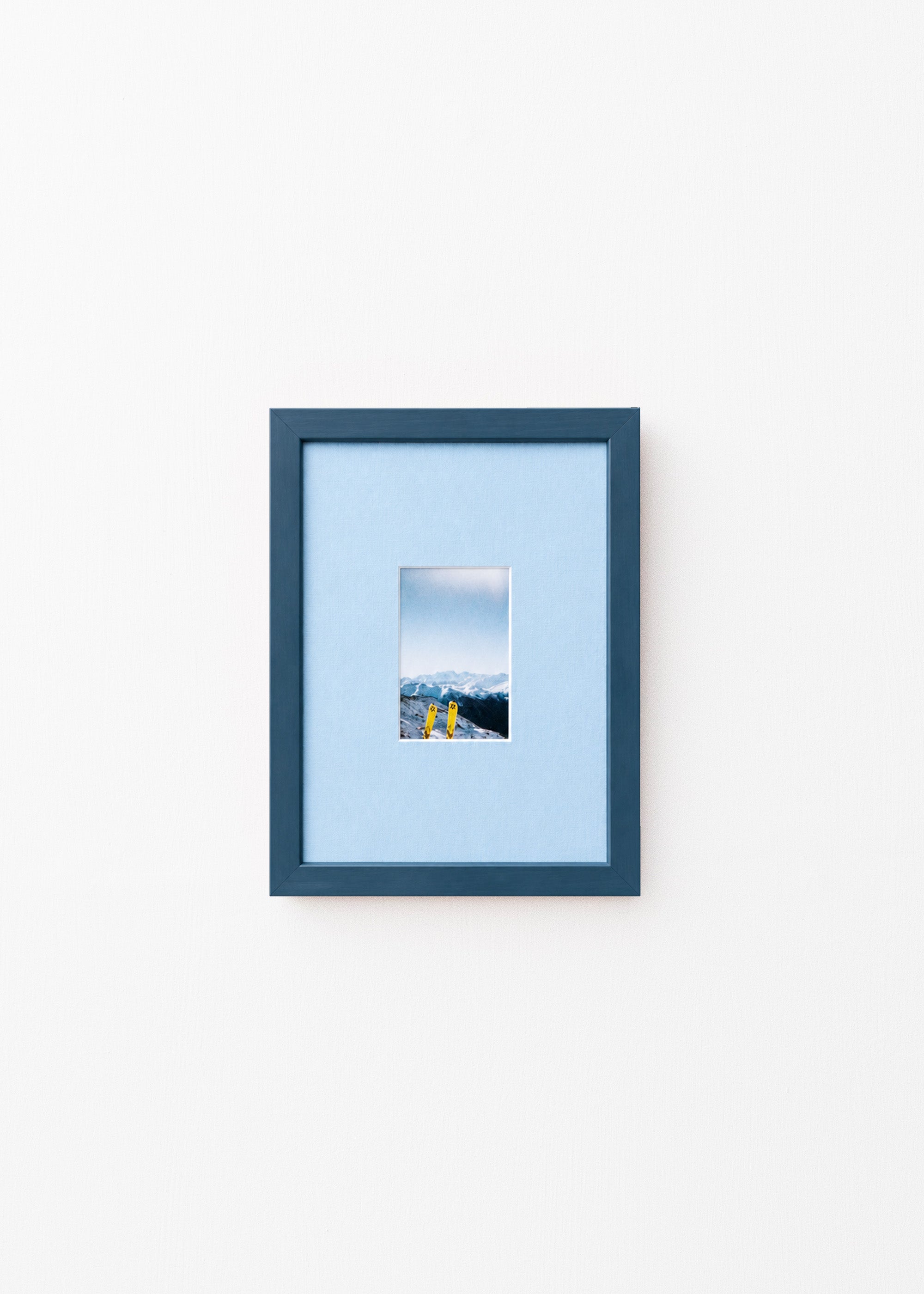 A dark blue picture frame with a light blue mat, displaying a small landscape photograph of mountains and yellow skis, set against a plain white wall.