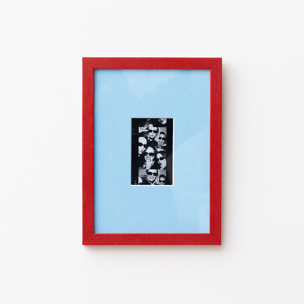 A framed art print featuring a black and white collage in a vertical black frame, set against a light blue mat and a vibrant red outer frame, mounted on a plain white wall.